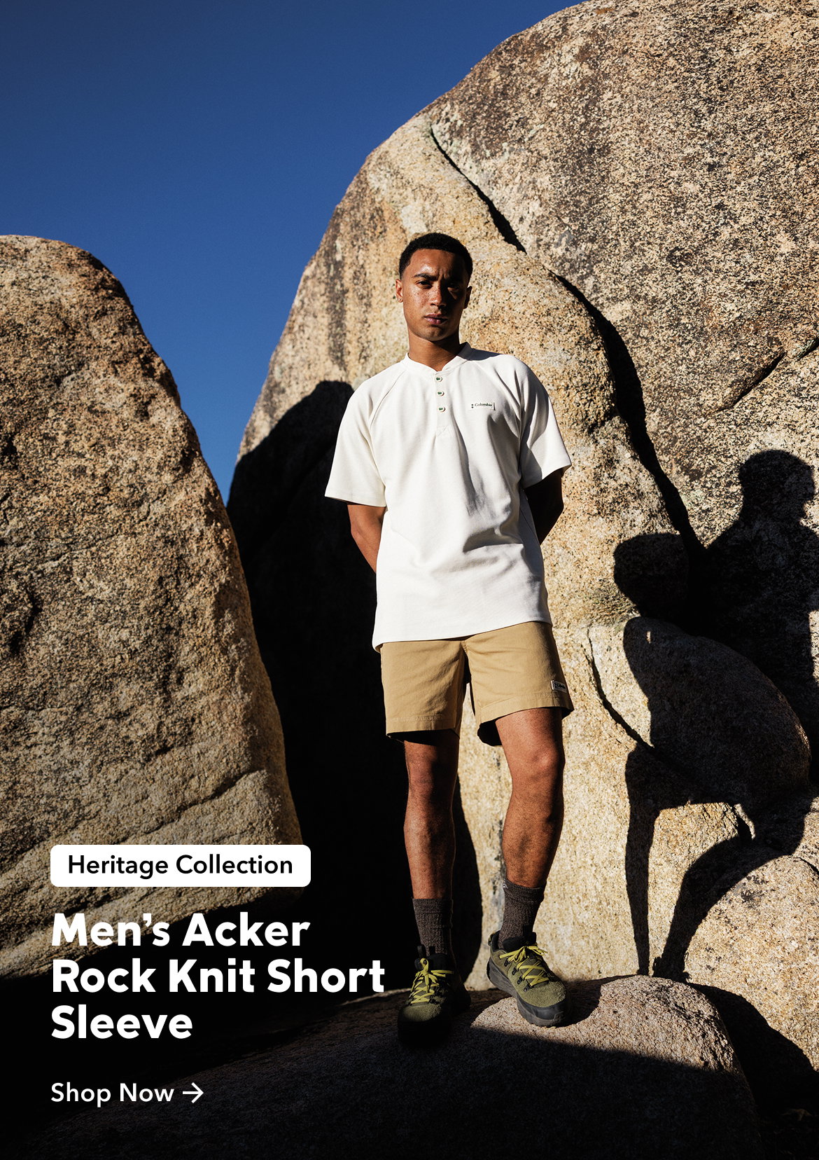 A man standing against a rock wearing a white short sleeve shirt, tan shorts, and green hiking boots with text: Heritage Collection Men's Acker Rock Knit Short Sleeve Shop Now