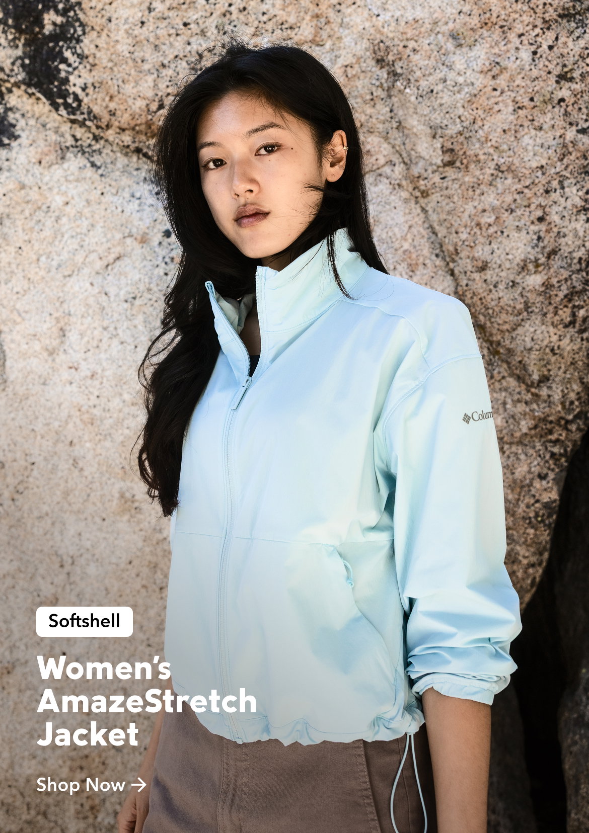 A woman standing against a rock wearing a blue zip-up jacket and mauve pants with text: Softshell Women's AmazeStretch Jacket Shop Now