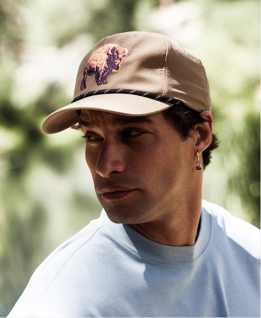 A man wearing a white crew neck t-shirt and a brown cap with a buffalo patch on the front, standing in the outdoors.