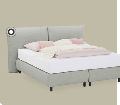 Ein hellgraues Boxspringbett mit rosafarbener und weißer Bettwäsche vor einem braunen Hintergrund.