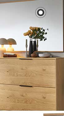 In der Mitte ist eine Kommode in Holzoptik mit schwarzen Elementen zu sehen. Darüber hängt ein rechteckiger Spiegel mit einer Lampe, einer Vase und weiteren Dekoelementen davor. Rechts hängt eine Garderobe in Holz mit einer hellen Jacke und einer braunen Tasche. Links befindet sich ein Schrank in Holzoptik mit schwarzen Elementen und ein großes Fenster mit Pflanzen und einem Hocker davor. 