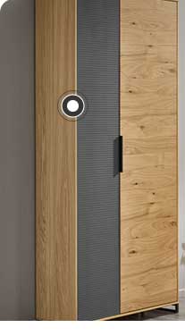 In der Mitte ist eine Kommode in Holzoptik mit schwarzen Elementen zu sehen. Darüber hängt ein rechteckiger Spiegel mit einer Lampe, einer Vase und weiteren Dekoelementen davor. Rechts hängt eine Garderobe in Holz mit einer hellen Jacke und einer braunen Tasche. Links befindet sich ein Schrank in Holzoptik mit schwarzen Elementen und ein großes Fenster mit Pflanzen und einem Hocker davor. 