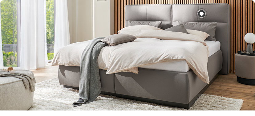 Ein graues Boxspringbett steht vor einer Wand mit Holzpanelen. Auf dem Bett liegt helle Bettwäsche und eine graue Kuscheldecke. Rechts neben dem Bett steht ein runder Beistelltisch mit einer Lampe. Links sieht man zwei bodentiefe Fenster mit hellen Gardinen und davor einen runden gepolsterten Hocker mit einer Decke. 