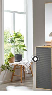 In der Mitte ist eine Kommode in Holzoptik mit schwarzen Elementen zu sehen. Darüber hängt ein rechteckiger Spiegel mit einer Lampe, einer Vase und weiteren Dekoelementen davor. Rechts hängt eine Garderobe in Holz mit einer hellen Jacke und einer braunen Tasche. Links befindet sich ein Schrank in Holzoptik mit schwarzen Elementen und ein großes Fenster mit Pflanzen und einem Hocker davor. 