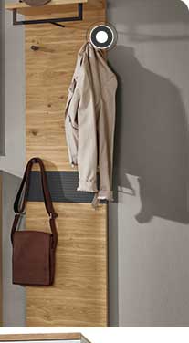 In der Mitte ist eine Kommode in Holzoptik mit schwarzen Elementen zu sehen. Darüber hängt ein rechteckiger Spiegel mit einer Lampe, einer Vase und weiteren Dekoelementen davor. Rechts hängt eine Garderobe in Holz mit einer hellen Jacke und einer braunen Tasche. Links befindet sich ein Schrank in Holzoptik mit schwarzen Elementen und ein großes Fenster mit Pflanzen und einem Hocker davor. 