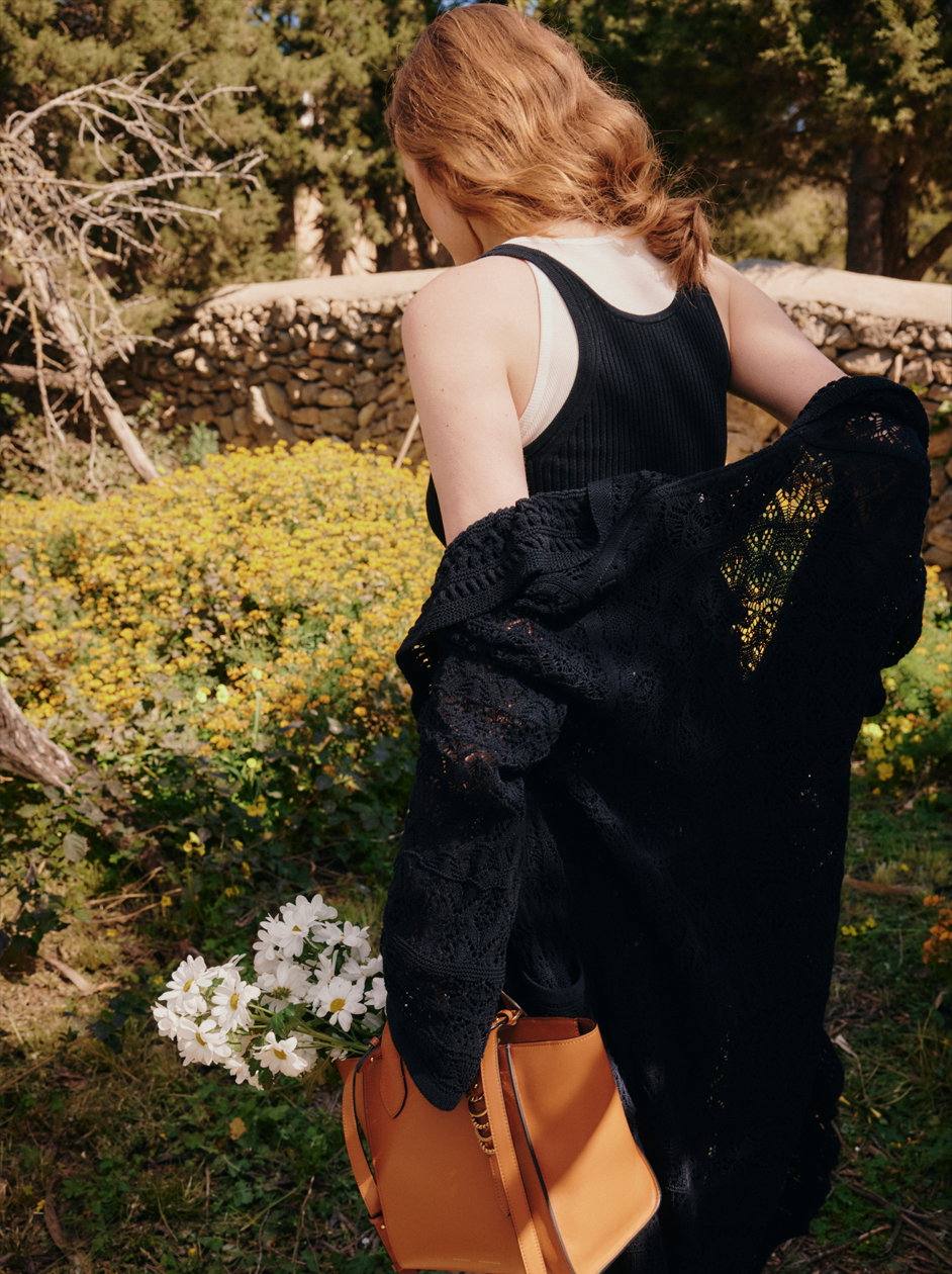 Ein Model von DOROTHEE SCHUMACHER in einer sonnigen Gartenlandschaft, von hinten gesehen. Sie trägt einen lässigen schwarzen Strick-Cardigan über einem schwarzen Ripp-Top und hält eine braune Ledertasche mit einem Strauß Margeriten in der Hand.