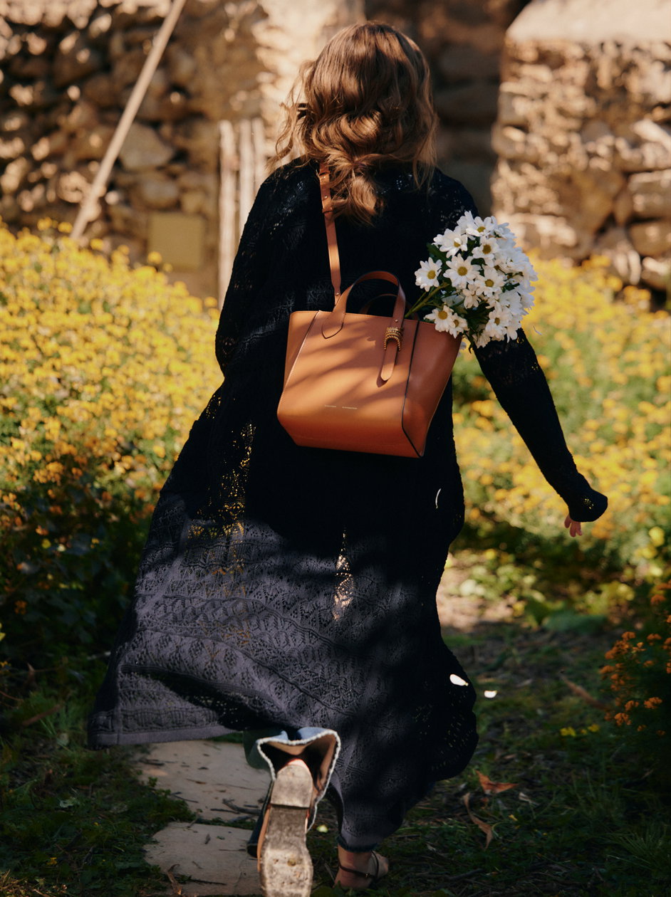 Rückenansicht eines Models von DOROTHEE SCHUMACHER, das in einem langen schwarzen Ajour-Strick-Cardigan durch ein Feld gelber Wildblumen läuft. Über der Schulter trägt sie eine cognacfarbene Ledertasche, aus der ein Strauß weißer Margeriten hervorschaut.