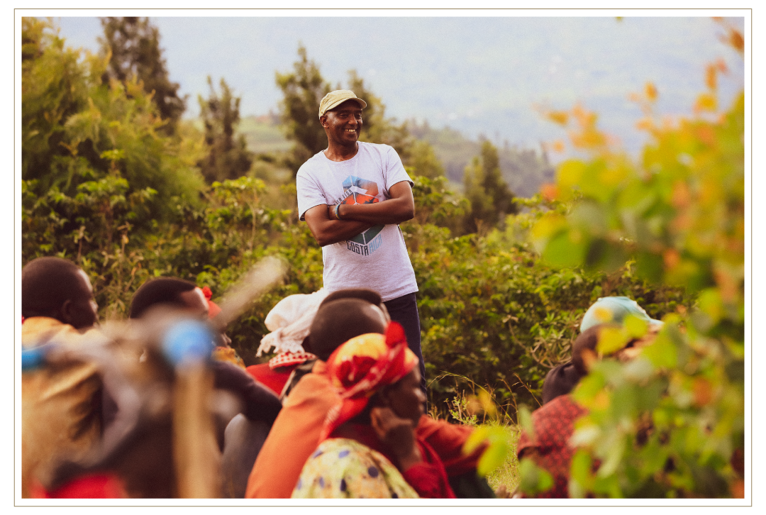 David Rubanzangabo stands outdoors at Huye Mountain with arms crossed, smiling as a group of people sit nearby, surrounded by lush greenery and a hillside landscape.