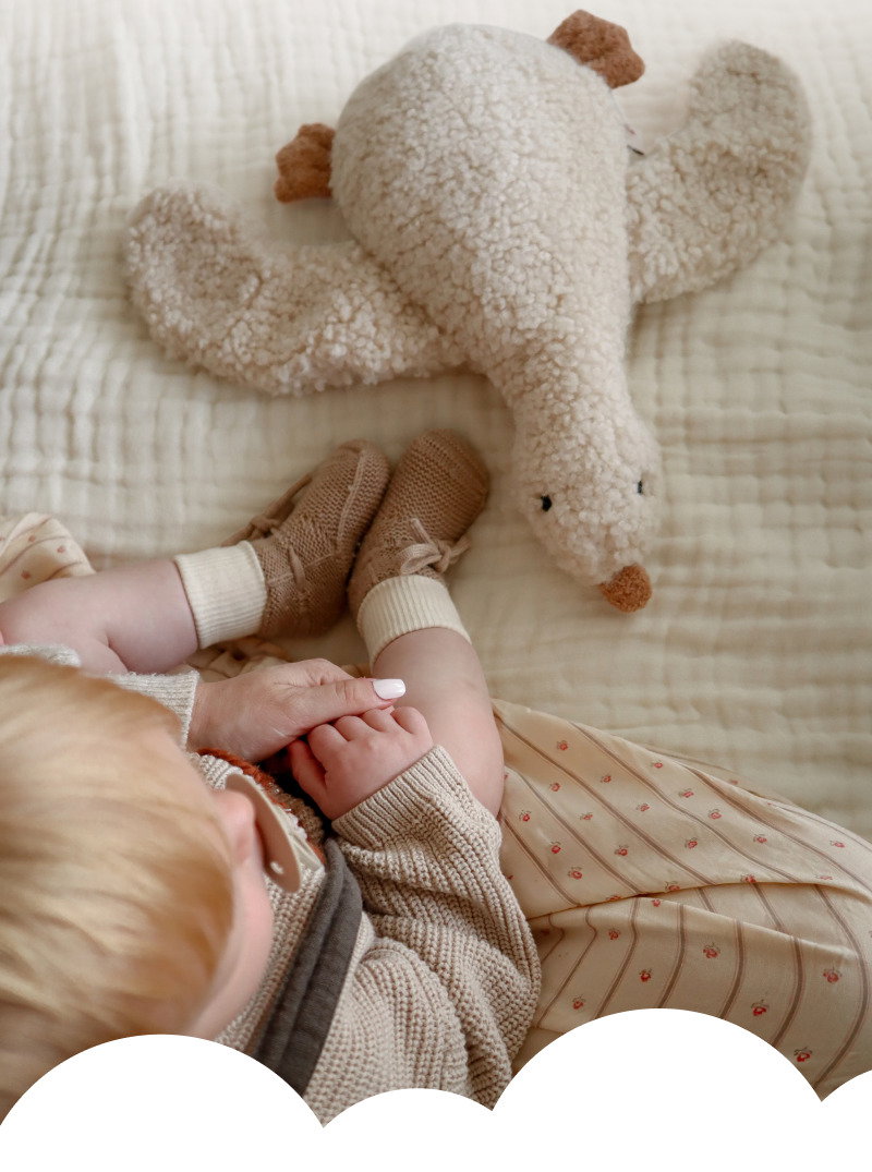Baby lying on a neutral blanket beside a large cream plush dog toy, with soft bedding and muted nursery tones.