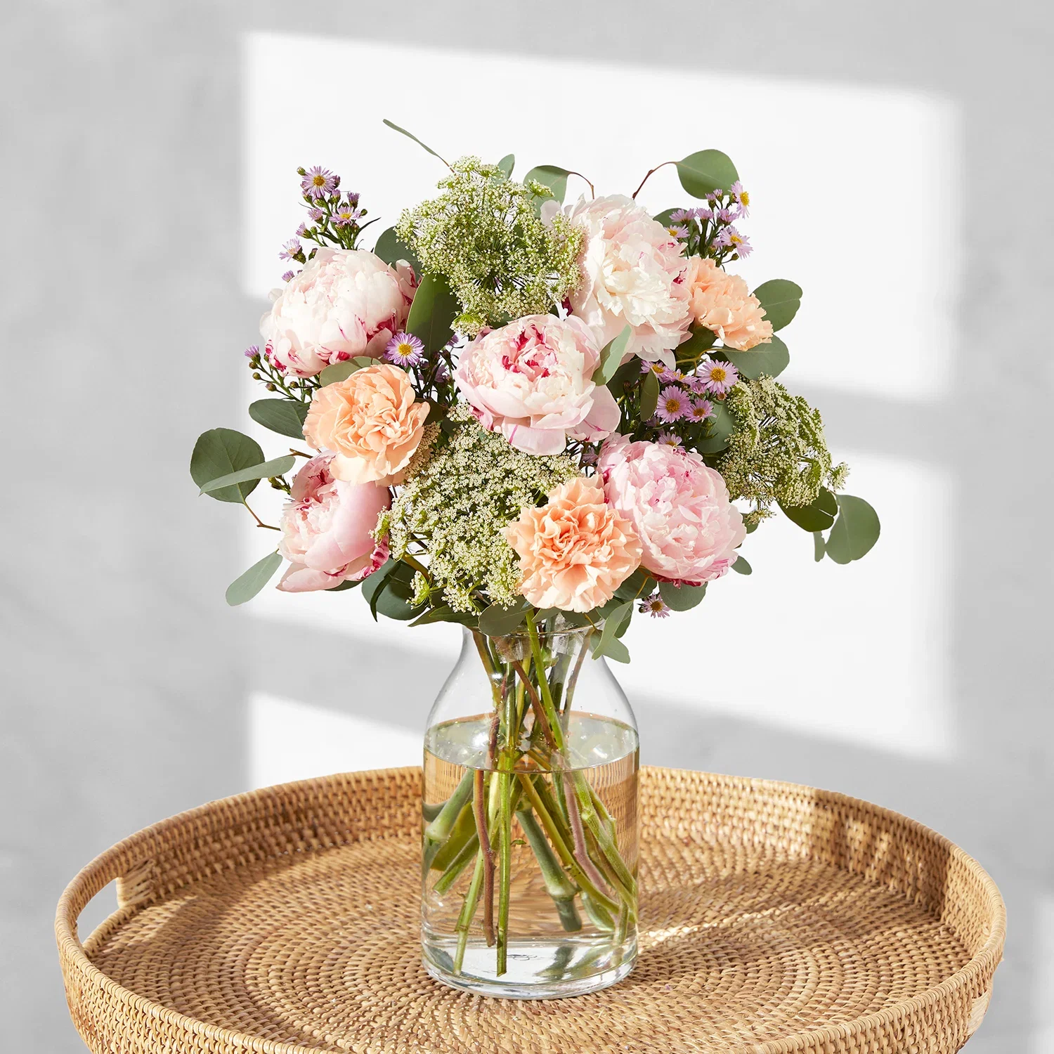 A delicate pastel bouquet styled in a clear glass jar vase on a woven rattan tray table. The arrangement displays layered, ruffled pale pink peonies, peach carnations, frothy clusters of white Ammi, and sprigs of tiny lilac aster daisies, interspersed with rounded green eucalyptus leaves against a sunlit background.