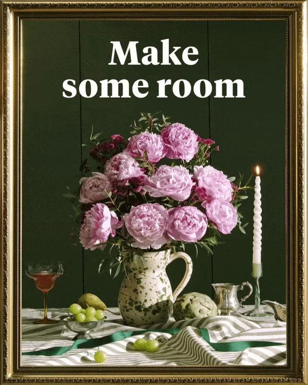 A decadent bouquet of large, ruffled pink peonies and dark purple accents in a green-speckled ceramic jug. The arrangement sits on a striped tablecloth alongside a lit twisted candle, a glass of rosé, and fresh fruit. Set against a dark green wall within a gold frame, text at the top reads ‘Make some room’. The camera slowly pans back to reveal the frame hanging.
