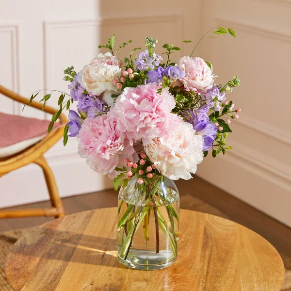 A soft, pastel floral bouquet presented in a clear glass vase on a wooden coffee table. The arrangement features large, fluffy pink and cream peonies, clusters of fragrant purple stocks, delicate bell-shaped freesia, and scattered pink hypericum berries with fresh green foliage.