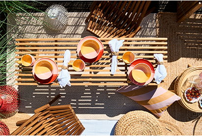 Draufsicht auf einen hölzernen Balkontisch, der im Boho-Stil mit gelb-rotem Geschirr, hellblauen Servietten und Gläsern gedeckt ist. Daneben sind geflochtene Hocker und gestreifte Kissen zu sehen. Rechts steht der Text ‚BOHO PICKNICK AUF BALKONIEN‘ mit einem Button ‚Zur Einrichtungsidee‘.