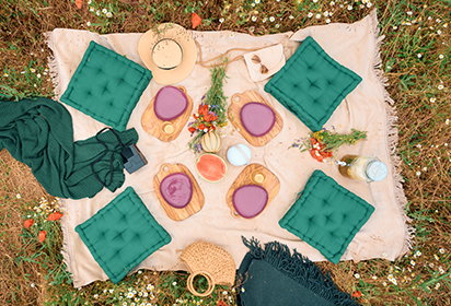 Vogelperspektive auf eine Picknickdecke auf einer Wiese, umgeben von grünen Sitzkissen. Auf der Decke sind hölzerne Servierbretter mit Snacks und bunten Tellern arrangiert. Rechts steht der Text ‚BRUNCH PICKNICK IM GRÜNEN‘ mit einem Button ‚Picknick entdecken‘.