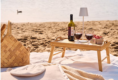 Ein kleines Holztablett steht auf einer Decke im Sand an einem Seeufer. Darauf befinden sich Weingläser, eine Flasche Wein, eine Schale Erdbeeren und eine moderne Akkuleuchte. Rechts steht der Text ‚SÜẞIGKEITEN-VASE MIT BLUMEN‘ mit einem Button ‚Zur Picknickidee‘.