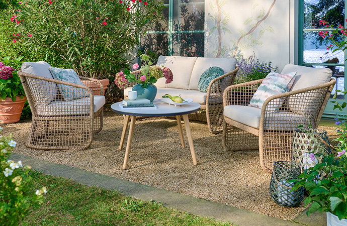 Eine gemütliche Sitzgruppe im Garten auf einer Kiesfläche: Bestehend aus einem Zweisitzer-Sofa und zwei Sesseln in heller Geflecht-Optik mit beigen Polstern. In der Mitte steht ein runder Holztisch mit einer blauen Blumenvase und Büchern; im Hintergrund blühen rote und rosa Hortensien.