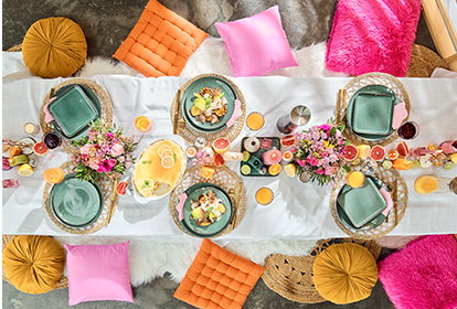 Draufsicht auf eine festlich gedeckte Picknicktafel am Boden mit vielen bunten Kissen in Pink, Orange und Gelb. Die Tafel ist mit Blumen, grünen Tellern auf Bast-Untersetzern und Früchten dekoriert. Rechts steht der Text ‚VERTRÄUMTE PICKNICKTAFEL‘ mit einem Button ‚Geschenkidee entdecken‘.