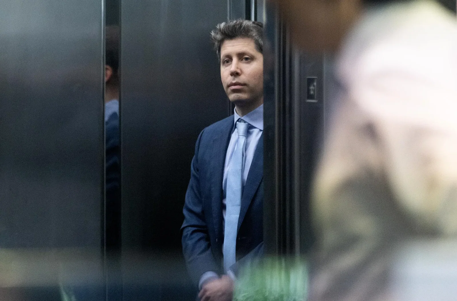 Sam Altman stands in an elevator, in a navy suit and light blue tie.