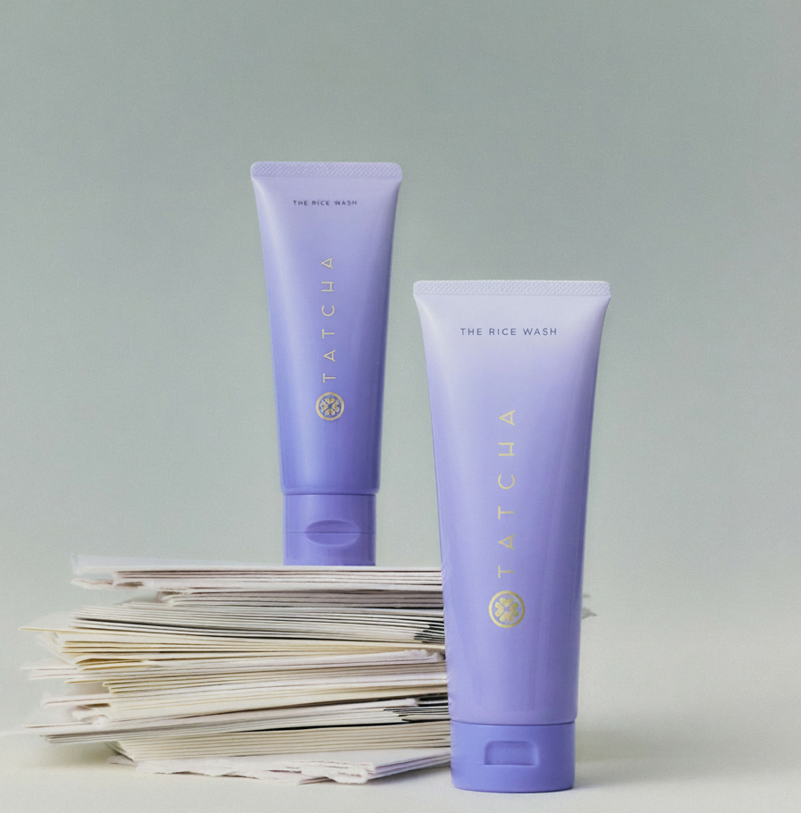 Two purple Tatcha The Rice Wash tubes stand on a stack of papers against a light green background.