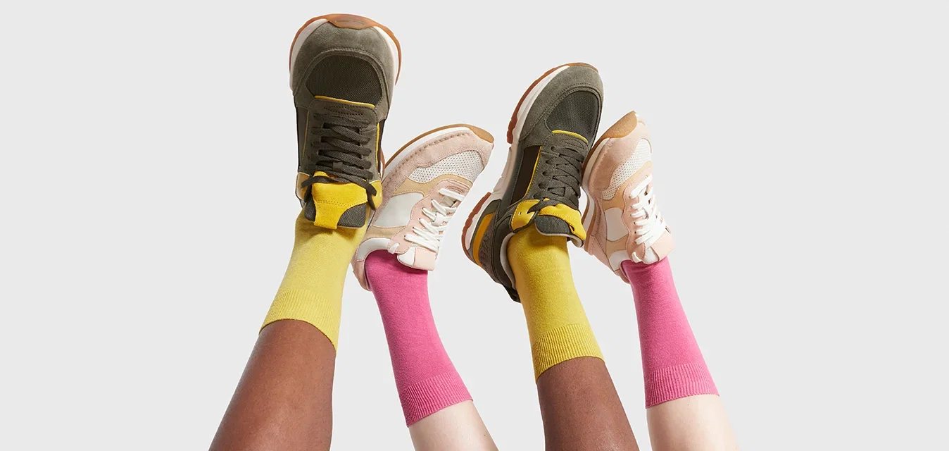 Twee paar sneakers: één olijfgroen met gele accenten, de andere beige met witte mesh. Heldere gele en roze sokken zichtbaar.