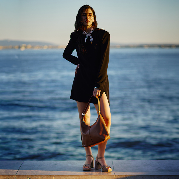 Woman in black dress holding brown handbag by the sea at sunset