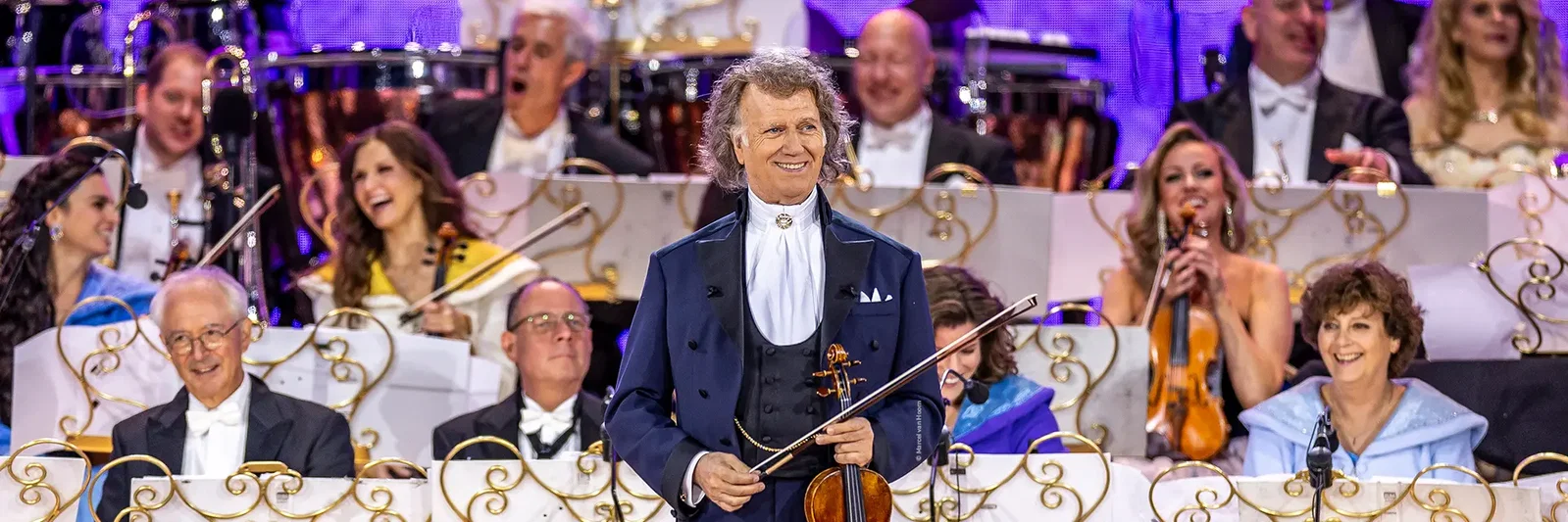 A conductor smiles while holding a violin, surrounded by an orchestra on stage.
