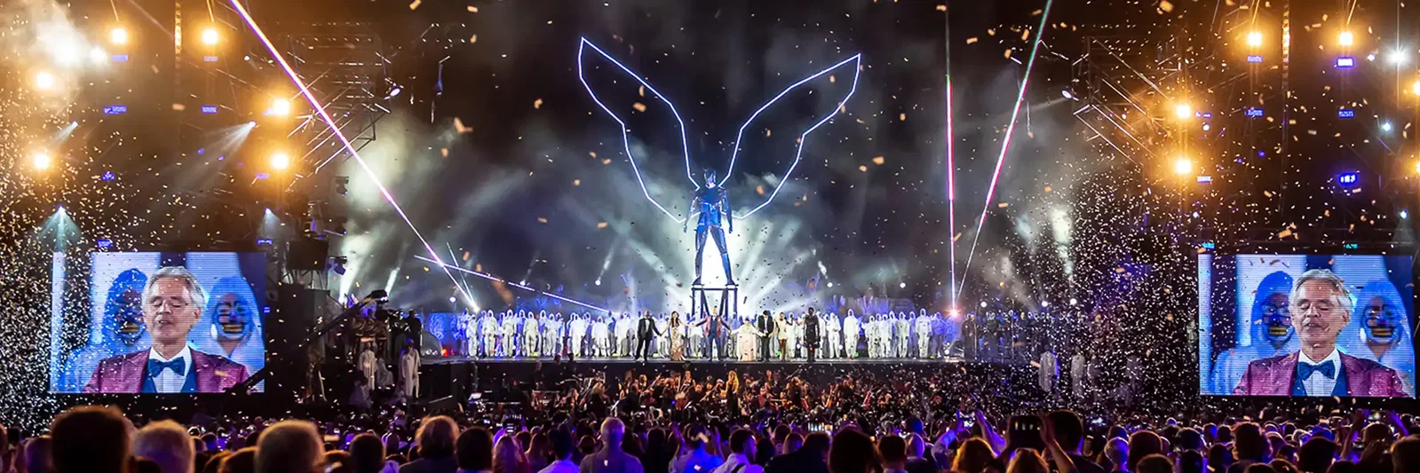 A vibrant stage performance with a large illuminated angel statue and a crowd celebrating.
