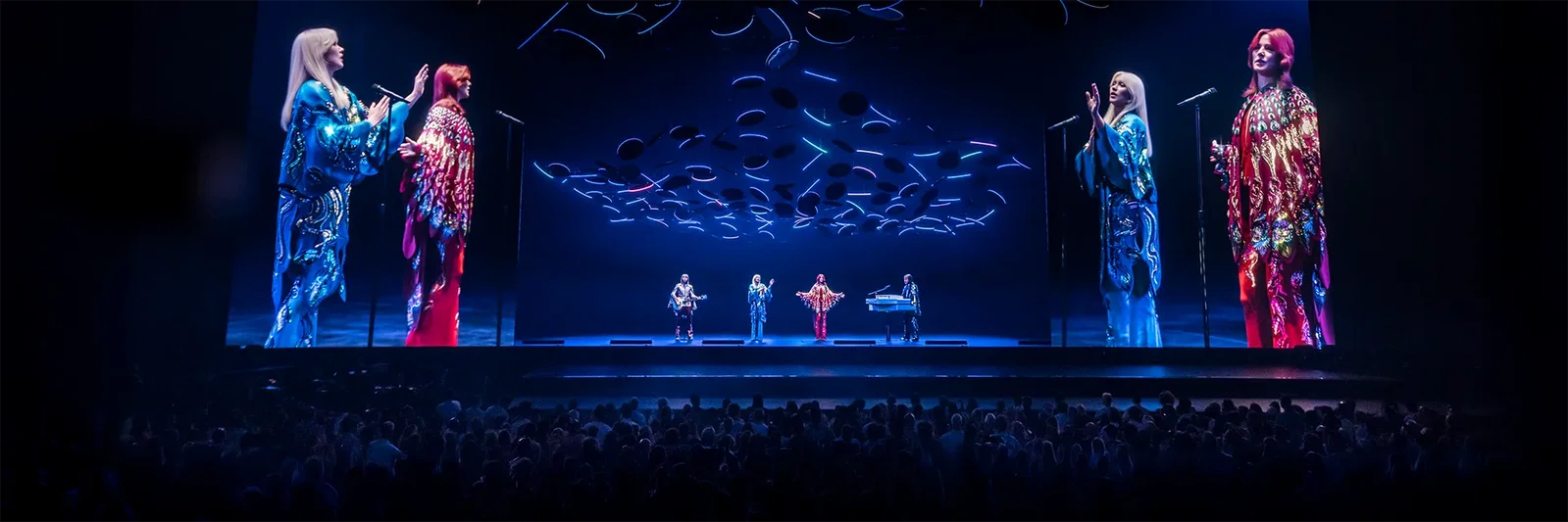 A vibrant stage performance with a large illuminated angel statue and a crowd celebrating.