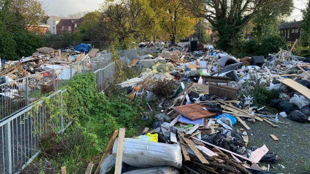 A huge amount of rubbish has been dumped close to a residential area, with houses and trees in the background.
