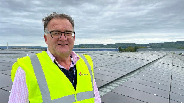 A man faces the camera smiling, wearing a pink shirt and a high-vis jacket. Behind him are hundreds of solar panels.