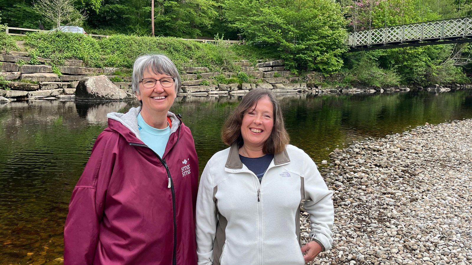 Di Leary and Karen Shackleton stand together in front of a bridge at the River Wharfe in Ilkley. They are standing on a pebbled shore with the brown but clear water of the river behind them.