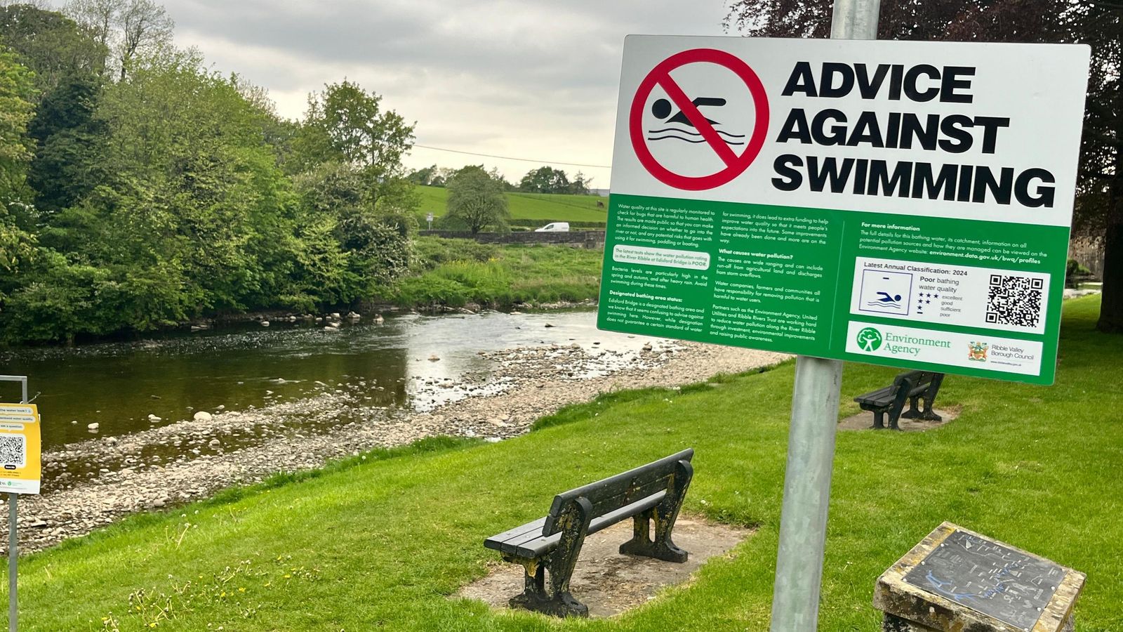 A sign saying Advice Against Swimming posted alongside a river in Lancashire. In the background is a river, alongside which are two benches.