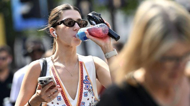 A woman drinks from a plastic bottle and holds a small hand-held fan. She has a mobile in her other hand. She is wearing a white vest top and sunglasses.