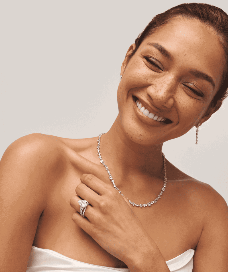 Smiling woman in bridal jewelry, including diamond drop earrings, diamond necklace and diamond pear-shaped engagement ring.