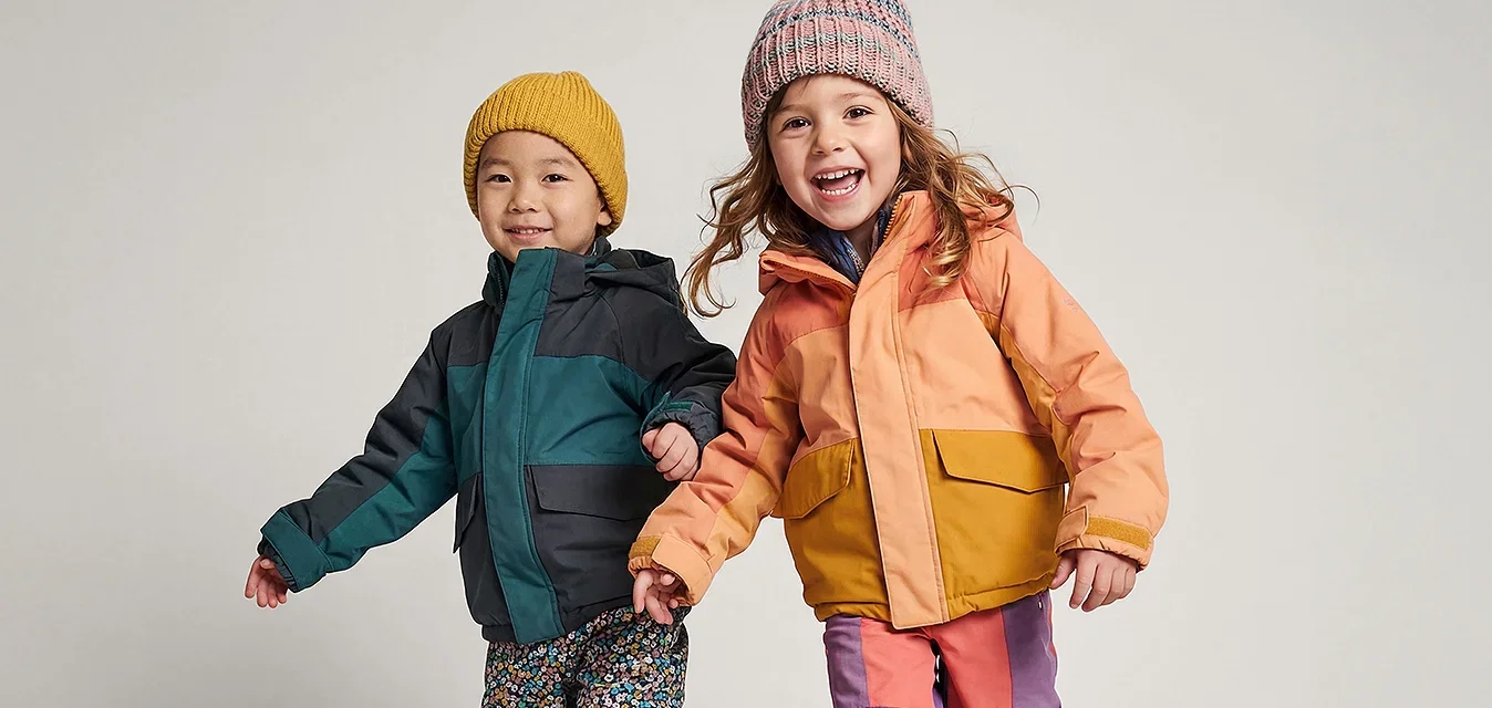 Zwei junge Kinder in bunten Winterjacken, Mützen und Stiefeln, die Händchen haltend vor einem schlichten Hintergrund nach vorne gehen.