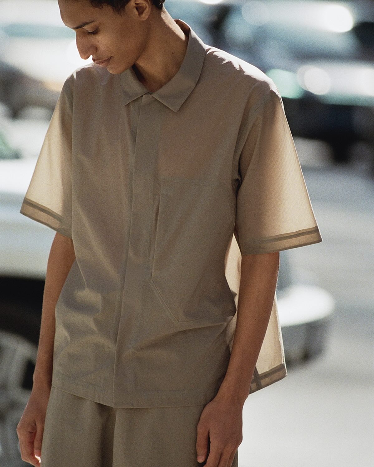 Young person wearing beige short-sleeve collared shirt and matching shorts, looking down on a blurred street
