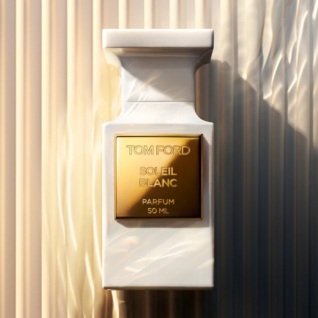 A white bottle of Tom Ford Soleil Blanc Parfum, 50 ml, with a gold label. The bottle is set against a textured, softly lit background, casting a subtle shadow.