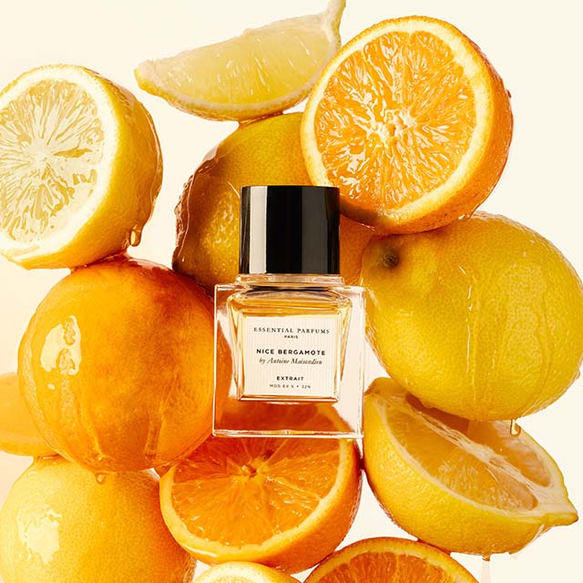 A bottle of Essential Parfums fragrance labelled 'Nice Bergamote' is surrounded by fresh citrus fruits including lemons, oranges, and a slice of grapefruit. The vibrant fruit arrangement highlights the perfume's citrus notes.