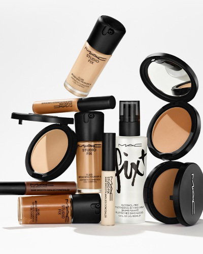 M.A.C cosmetic products including foundation bottles, a setting spray, and powder compacts are arranged aesthetically against a plain background. Some compacts are open, revealing their mirrors and shades.