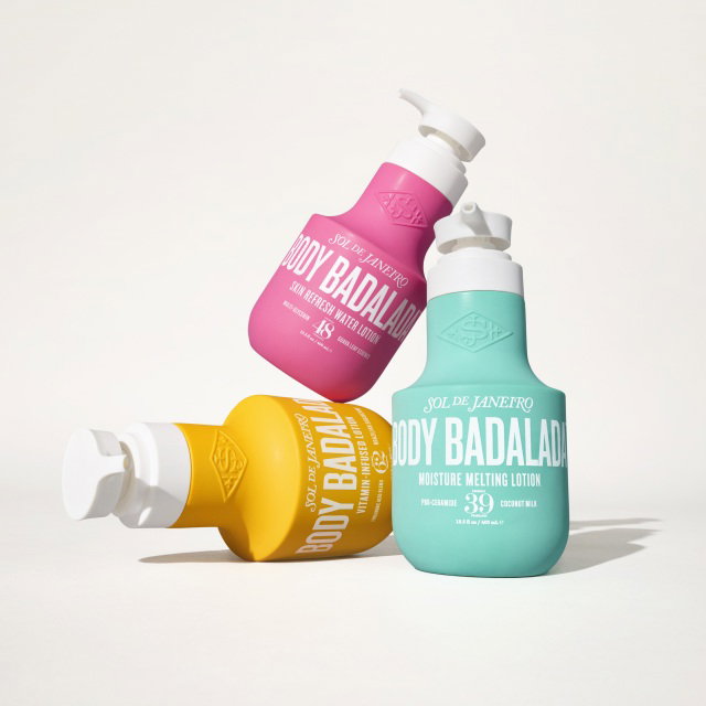 Three bottles of Sol de Janeiro Body Badalada Moisture Melting Lotion are arranged artistically. The bottles are in pastel shades of pink, yellow, and teal with white pump tops, showcasing the brand's logo and product details on the front. The backgr