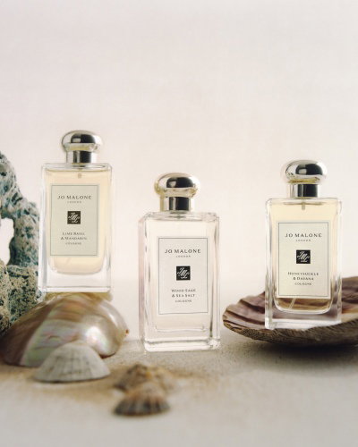 Three Jo Malone London perfume bottles are arranged on a sandy surface, surrounded by seashells and coral. Each bottle has a sleek design with a metallic cap, and the labels display the fragrance names. The composition evokes a coastal or beach theme