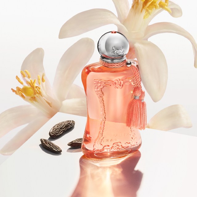 A Parfums de Marly perfume bottle with a pink liquid and a silver cap, adorned with a decorative tassel. The bottle is set against a white background with white flowers and tonka beans nearby, creating an elegant and luxurious presentation.
