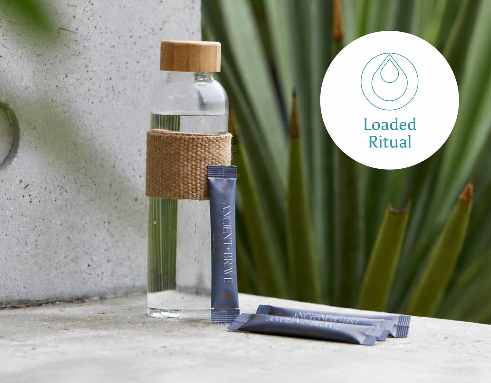A glass water bottle with a bamboo lid and woven jute sleeve sits on a stone ledge beside three navy Ancient + Brave sachets, with a large agave plant in the background. Loaded Ritual.
