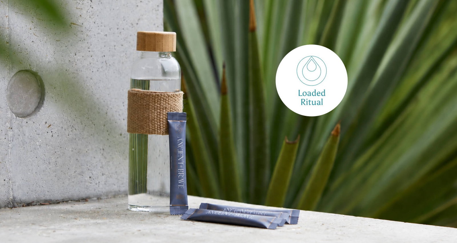 Here's an alt text for this one: A glass water bottle with a bamboo lid and woven jute sleeve sits on a stone ledge beside three navy Ancient + Brave sachets, with a large agave plant in the background. Loaded Ritual.