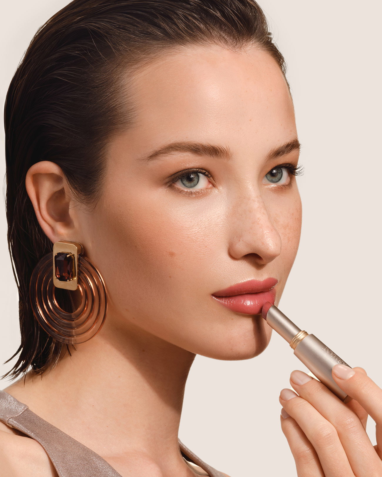 Primer plano de belleza de una modelo sosteniendo un stick plateado de Armani beauty cerca del rostro, sobre un fondo neutro claro. La modelo lleva un gran pendiente escultórico y un rectángulo beige grande cubre la parte central del rostro.