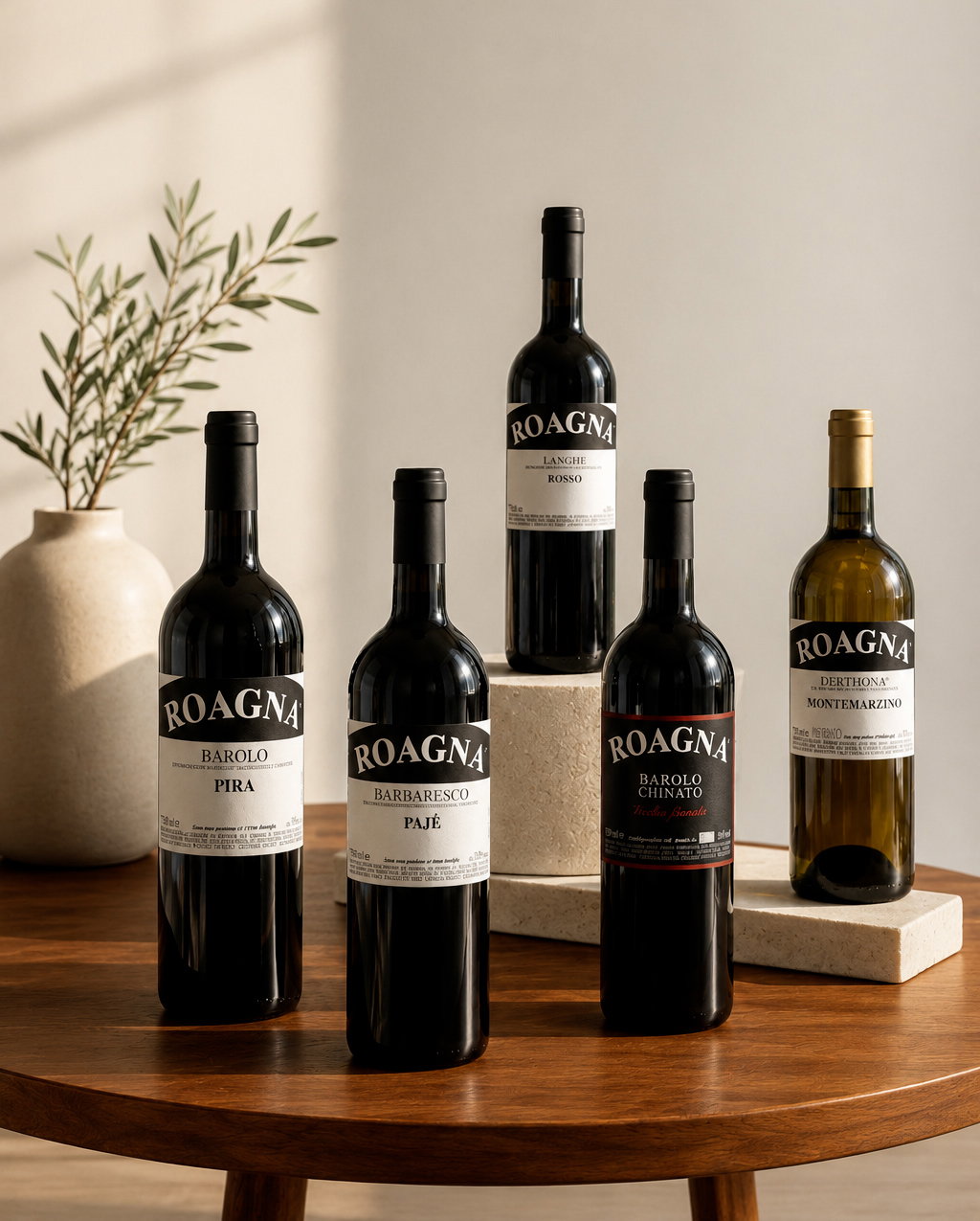 Five Roagna wine bottles (Barolo, Barbaresco, Langhe Rosso, Derthona) displayed on a wooden table with a ceramic vase and neutral backdrop