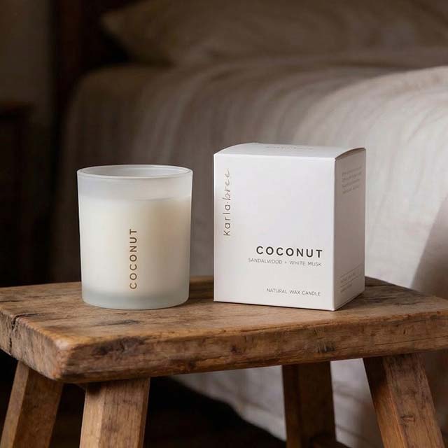 A Karla Bree coconut-scented candle in a glass holder sits beside its box on a wooden stool.