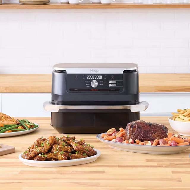 A Ninja appliance on a countertop surrounded by plates of food, including meats and fries.