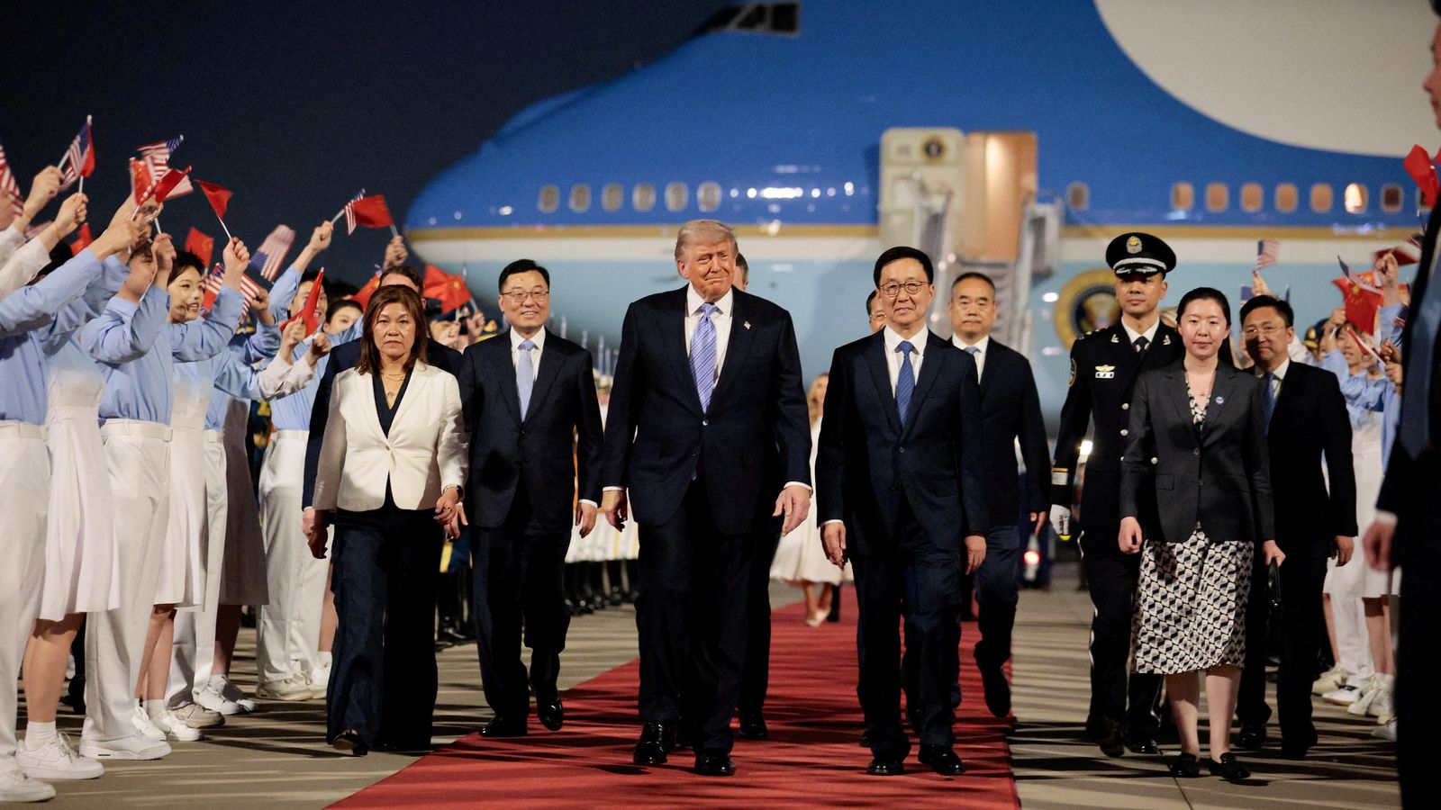 Donald Trump walks next to Chinese officials on a red carpet with people waving flags and a blue and white uniform lined up in rows either side of the walkway. Air Force One is in the background.