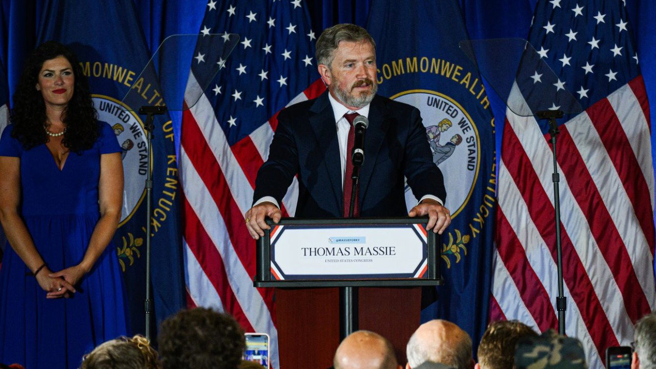 Thomas Massie stands behind a podium with his name on in front of a stage backdrop featuring US flags. There are rows of people in a crowd below the stage facing him and a woman to the left. He wears a dark suit with a red tie.
